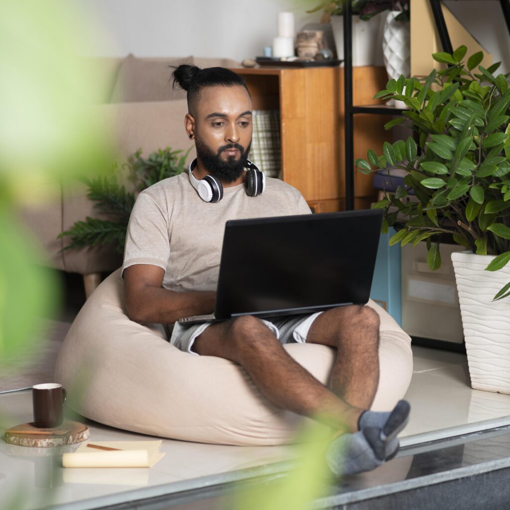 A casually dressed man with a beard and a top knot sits on a beige beanbag chair in a plant-filled living room, focusing on a black laptop with headphones resting around his neck.